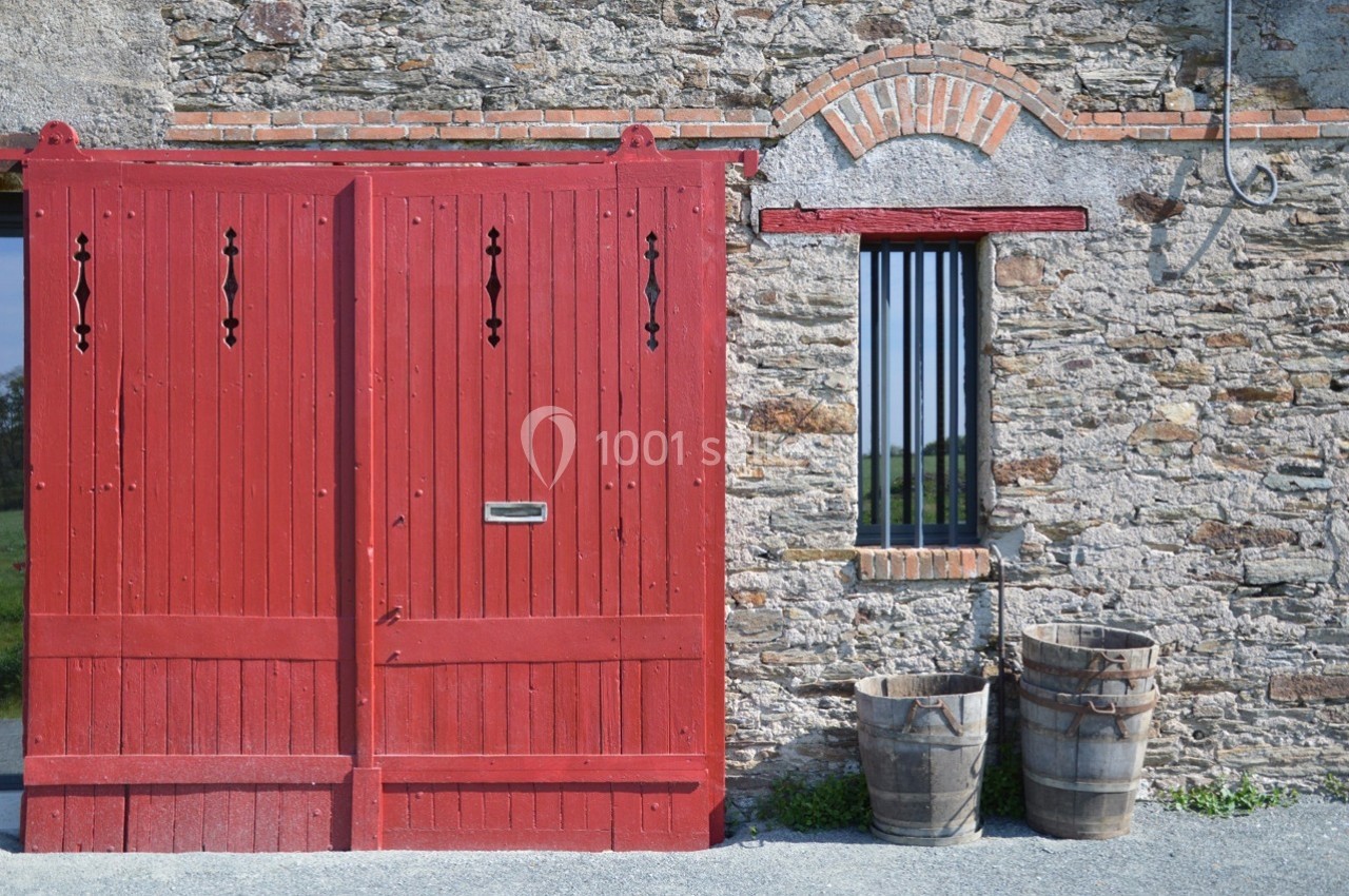 Porte en bois rouge et fenêtre à barreaux sur un mur en pierre, avec deux tonneaux en bois au premier plan.