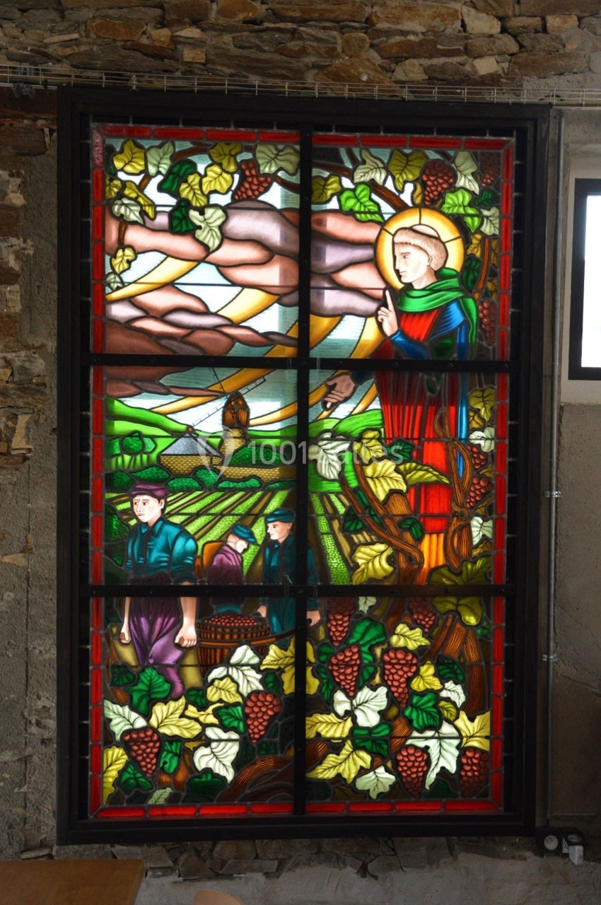 Vitrail coloré représentant une scène rurale avec des vignes, des travailleurs et un personnage religieux.