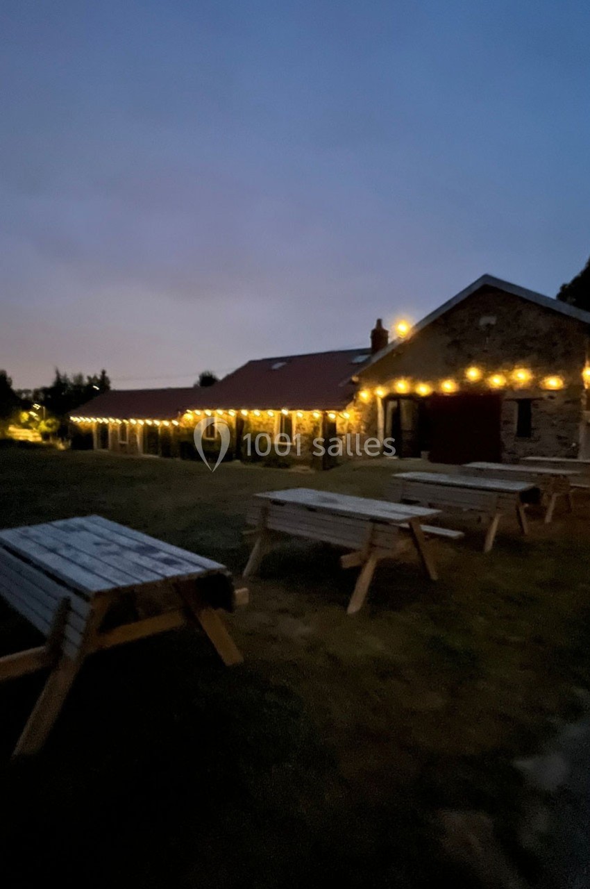 Bâtiment en pierre éclairé par des guirlandes lumineuses au crépuscule, avec des tables en bois sur une pelouse.