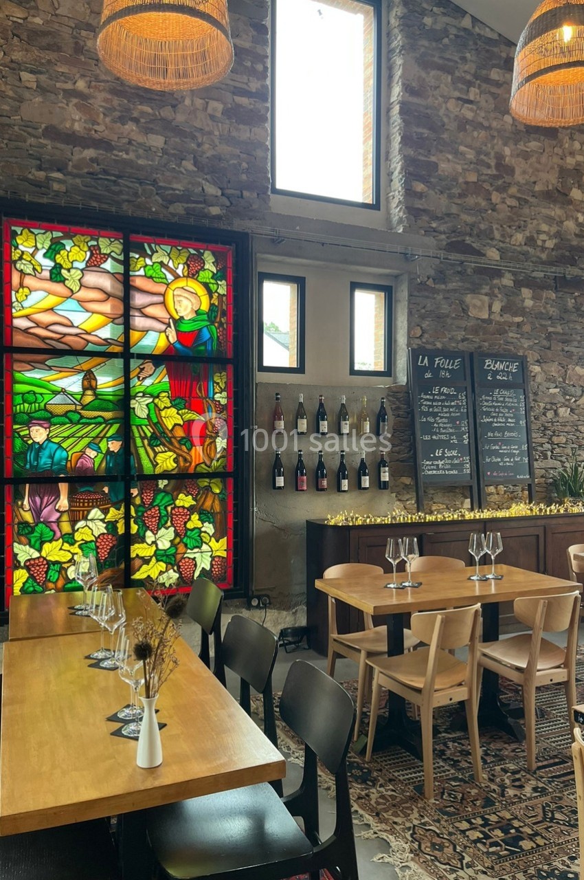 Salle de restaurant avec tables en bois, vitrail coloré, étagère à bouteilles et ardoises de menu sur un mur en pierre.