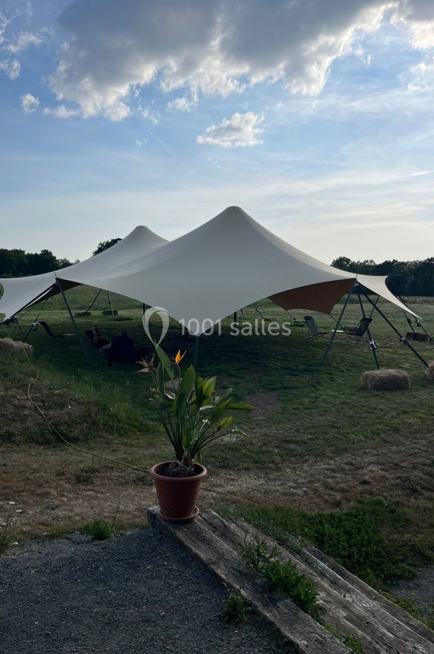 Tente blanche tendue sur une pelouse, entourée de bottes de foin, avec un pot de fleurs au premier plan.