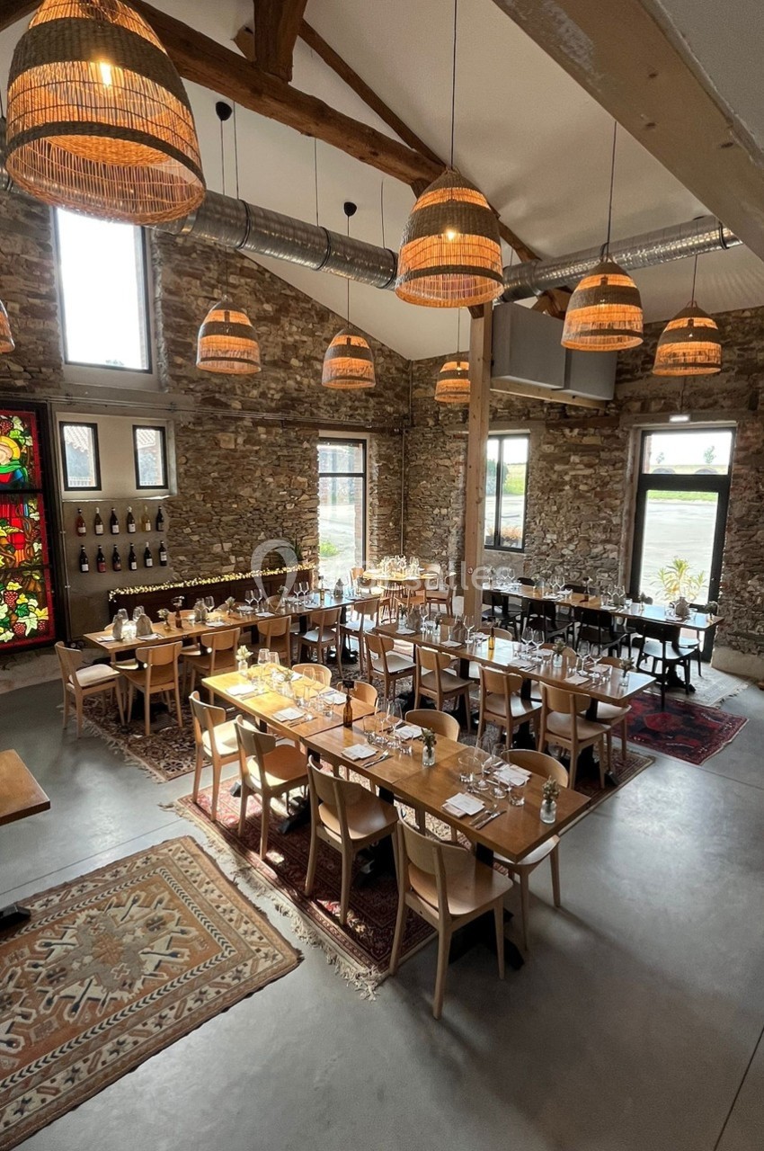 Salle de réception lumineuse avec murs en pierre, grandes fenêtres, tables dressées et suspensions en osier.