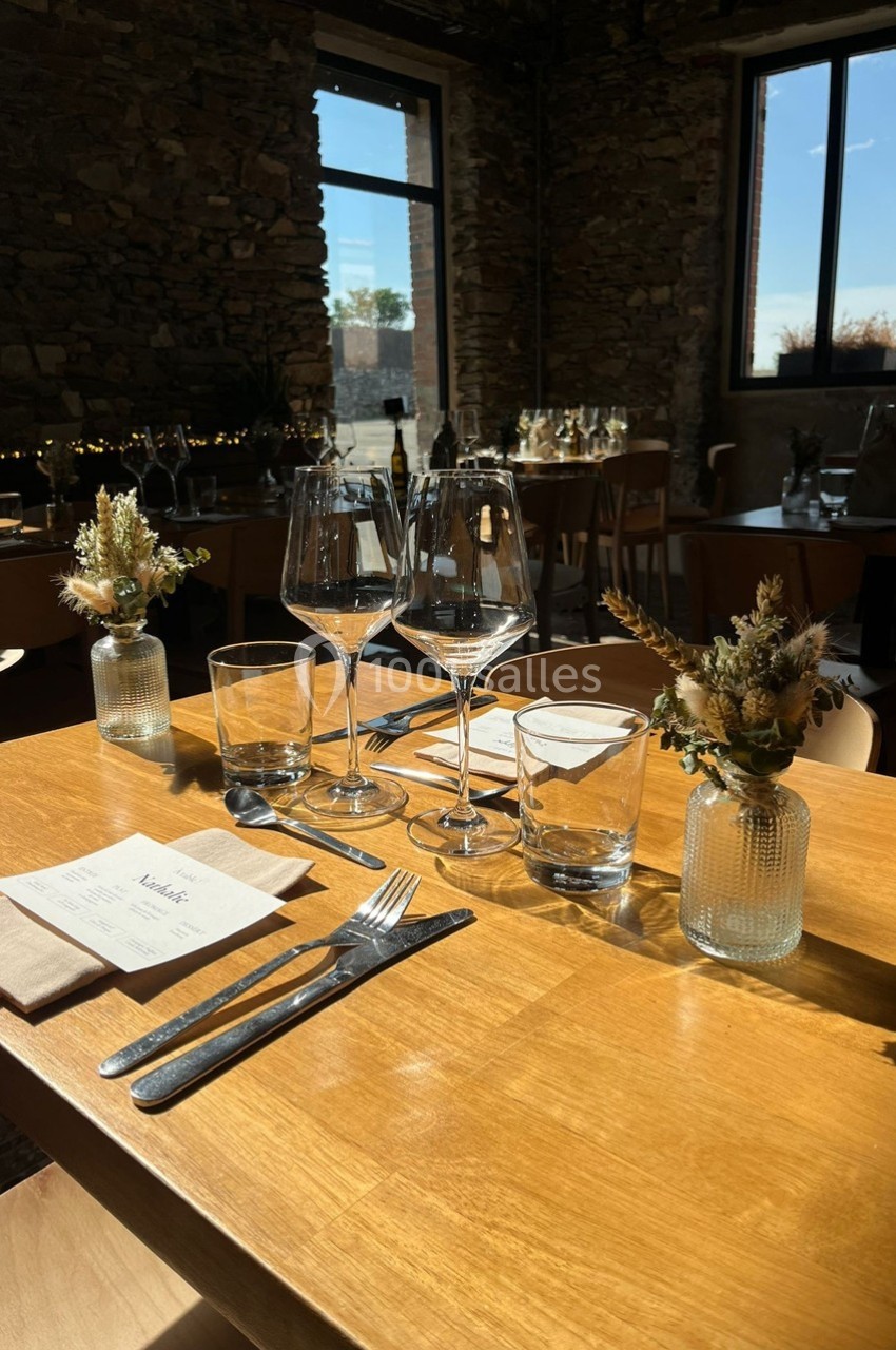Table en bois dressée avec verres, couverts et décorations florales, dans une salle lumineuse aux murs en pierre.