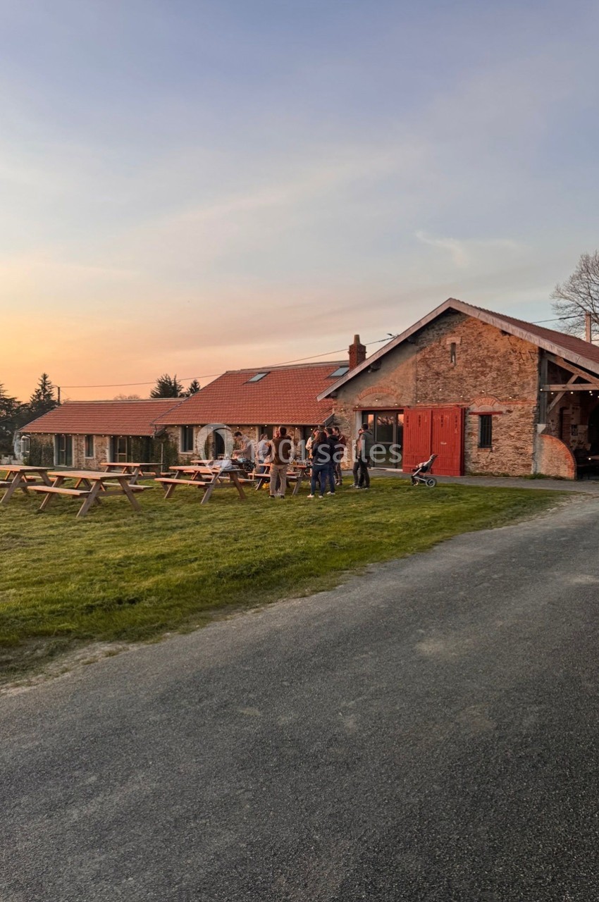 Groupe de personnes discutant devant un bâtiment en pierre avec toit en tuiles, entouré de tables de pique-nique au coucher…