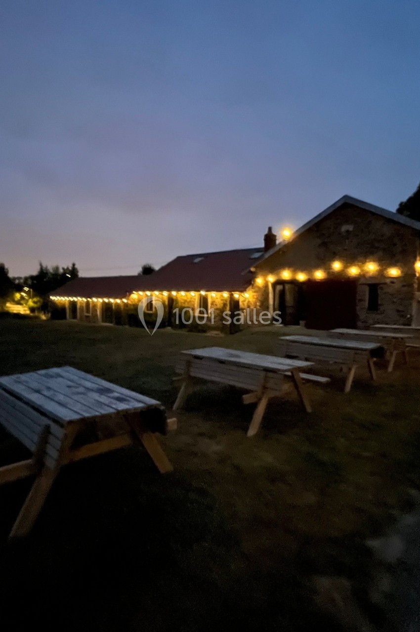 Bâtiment en pierre éclairé par des guirlandes lumineuses, avec des tables en bois sur une pelouse au crépuscule.