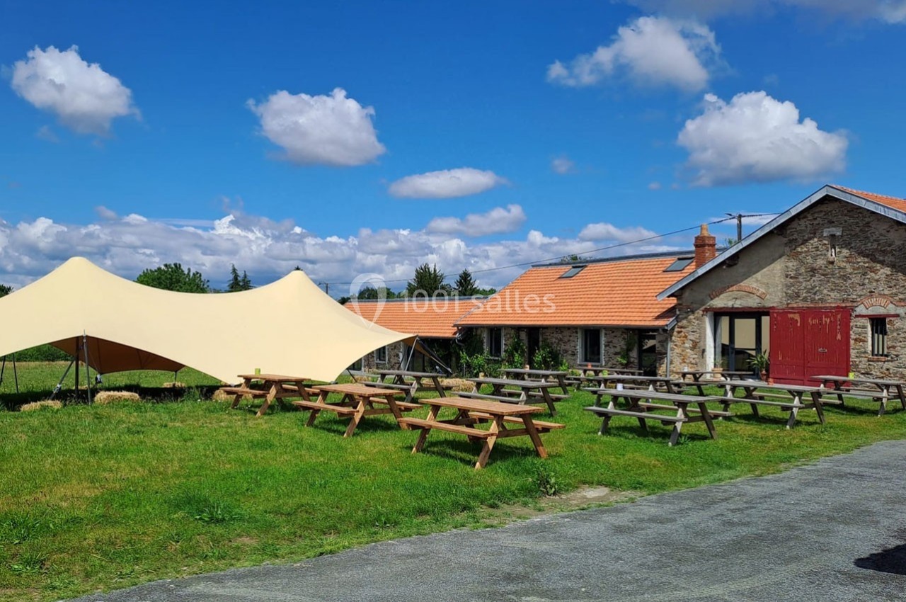 Bâtiment en pierre avec toit en tuiles, tables de pique-nique en bois et tente beige dans un espace vert sous un ciel bleu.
