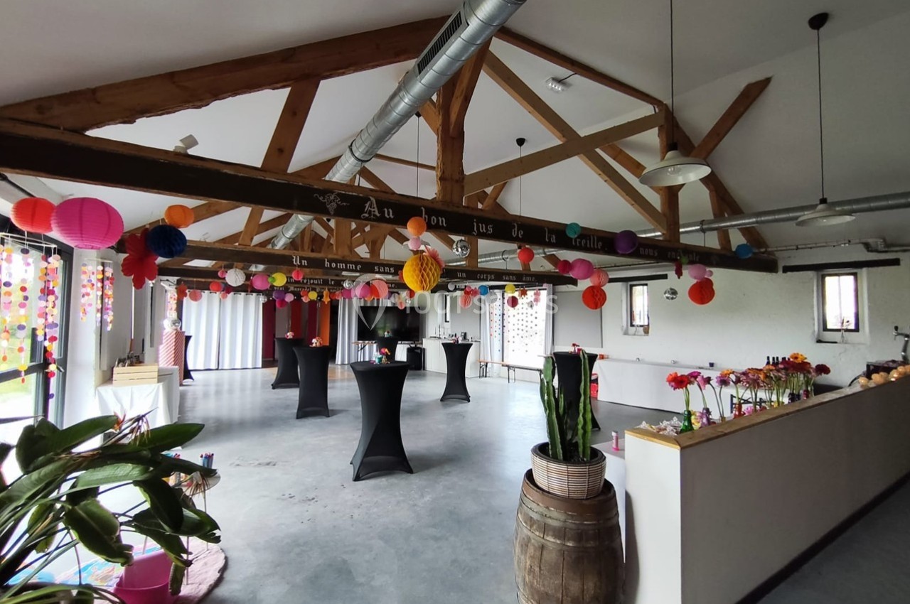 Salle décorée pour un événement avec guirlandes colorées, tables hautes noires et plantes en intérieur lumineux.