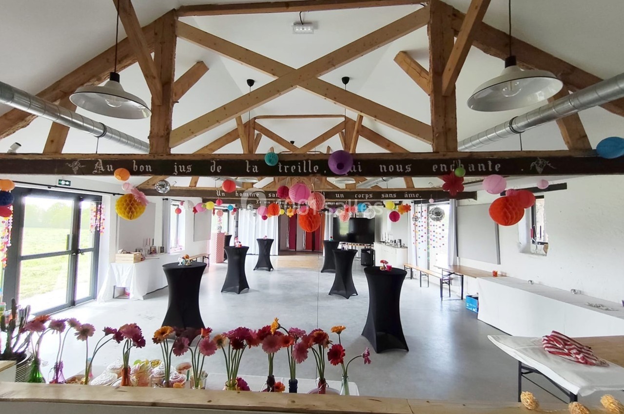 Salle décorée pour un événement, avec guirlandes colorées, fleurs et tables hautes sous une charpente en bois.