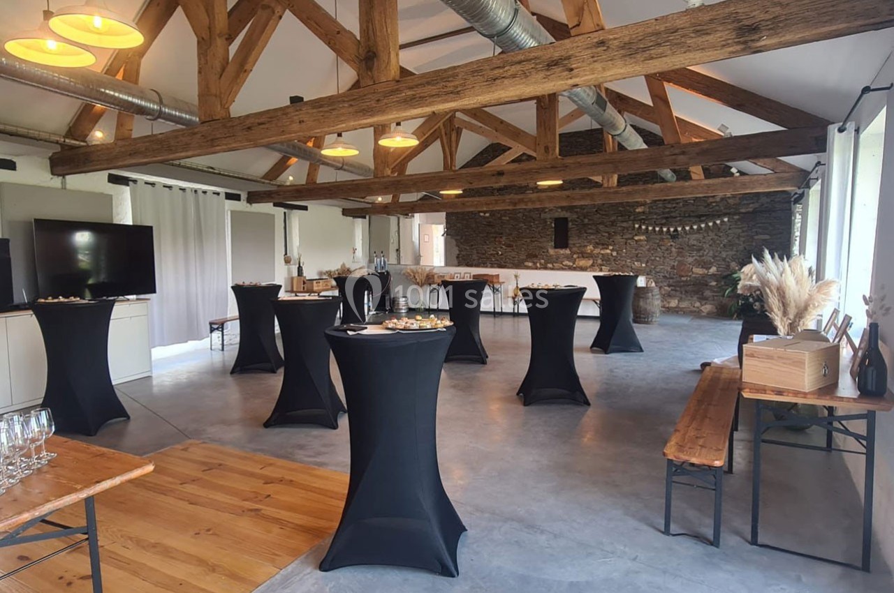 Salle de réception avec poutres apparentes, tables hautes noires et buffet disposé le long d'un mur en pierre.