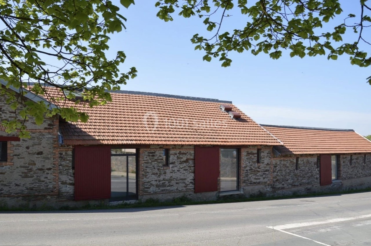 Bâtiment en pierre avec toit en tuiles rouges, grandes portes rouges et fenêtres, bordé par une route et des arbres.