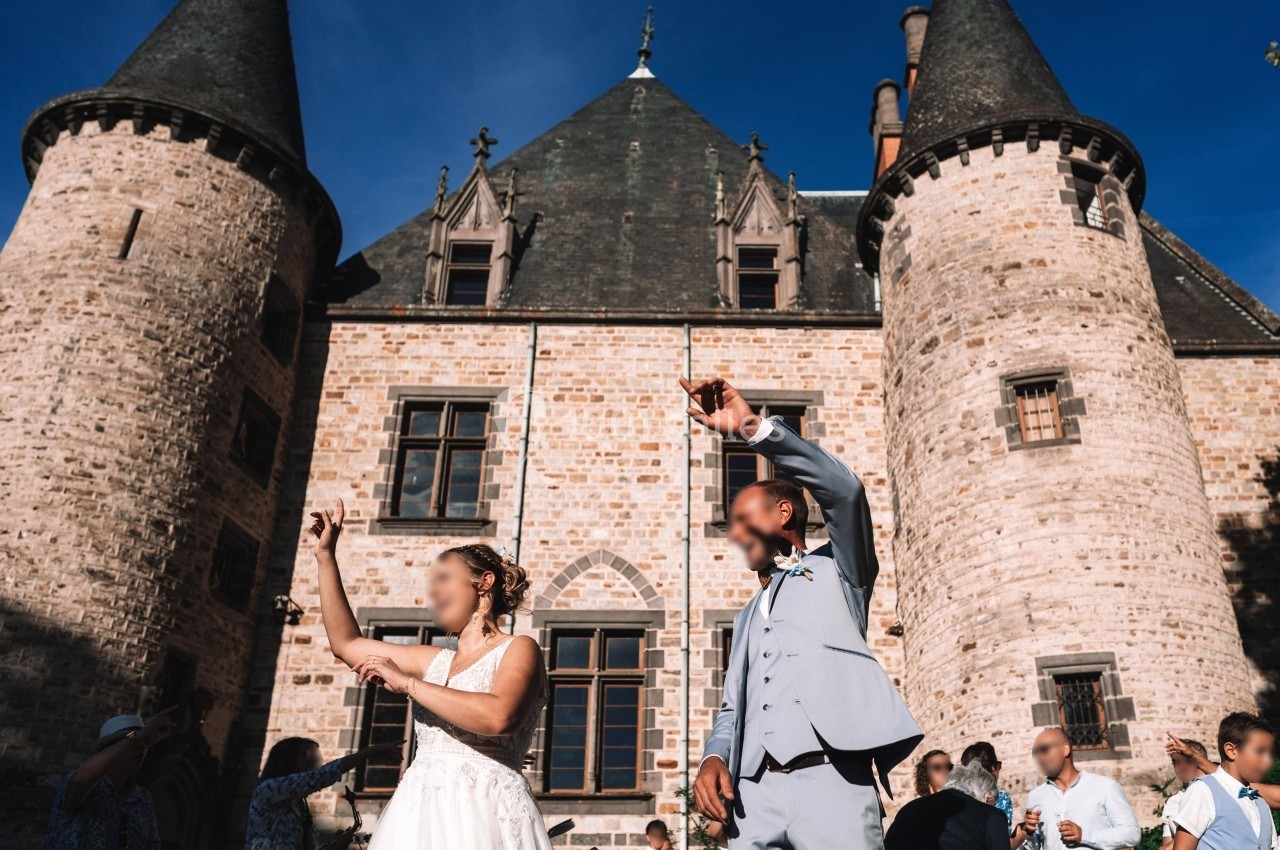 Un couple danse devant un château en pierre sous un ciel bleu, entouré de plusieurs personnes.