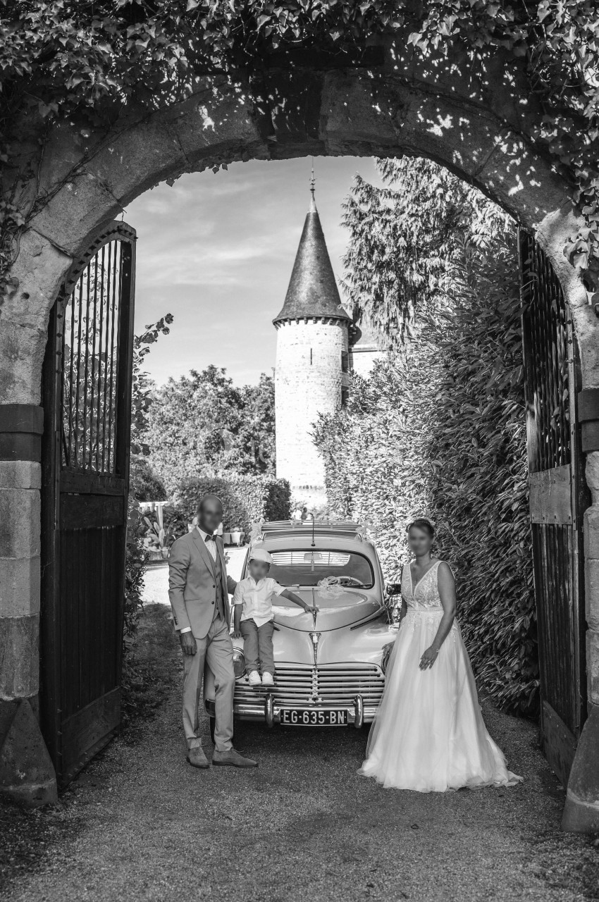 Un couple en tenue de mariage pose avec un enfant devant une voiture ancienne sous une arche, près d'une tour.