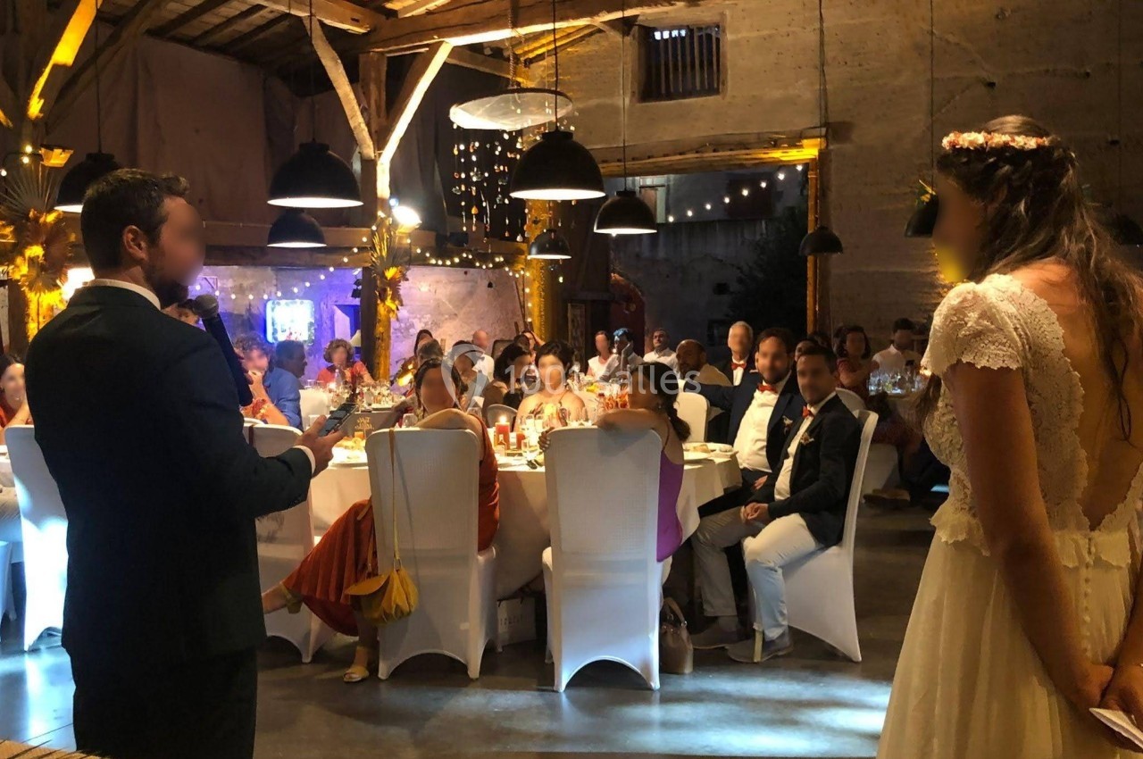 Un marié fait un discours devant des invités assis dans une salle de réception décorée, la mariée l'écoute.