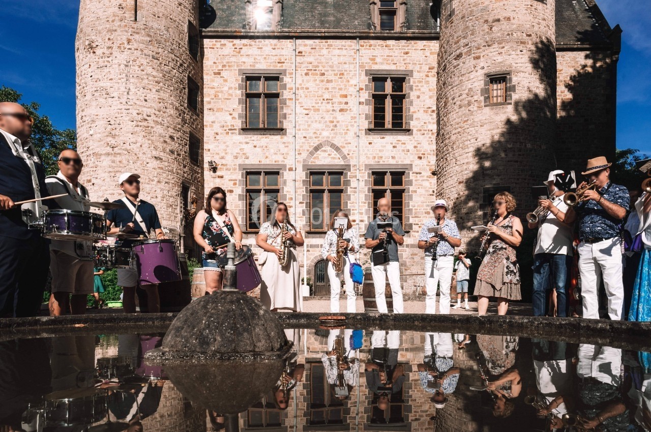 Un groupe de musiciens et de spectateurs devant un château en pierre, avec leur reflet visible dans une flaque d'eau.