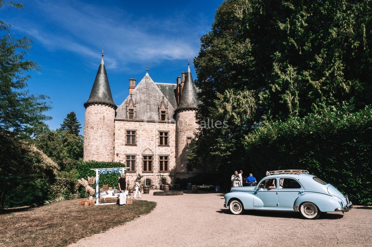 Château en pierre avec tours, entouré de végétation, une voiture ancienne bleue garée au premier plan.