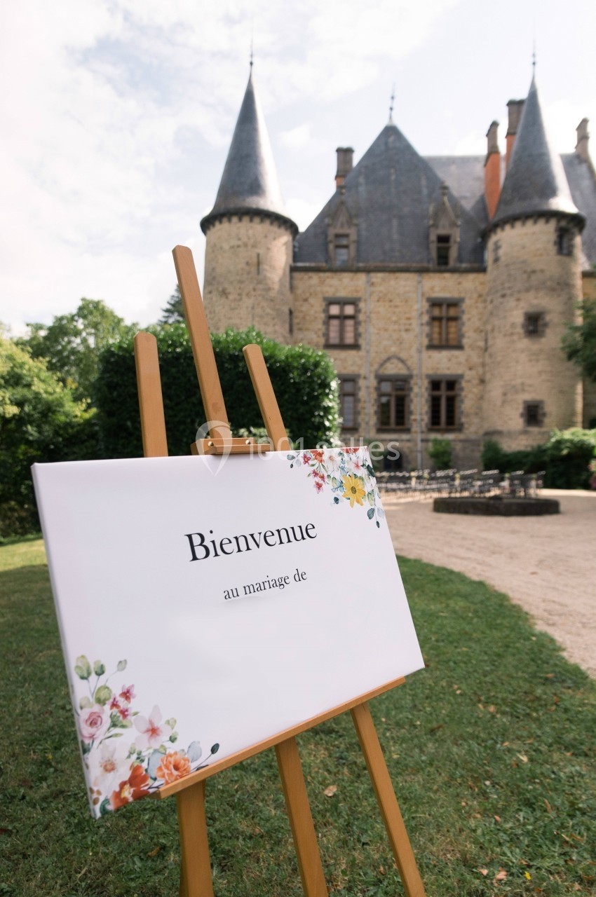 Panneau de bienvenue décoré de fleurs devant un château en pierre entouré de verdure.