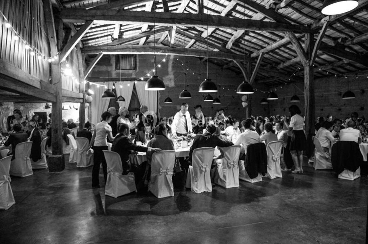 Salle de réception en bois avec des invités assis à des tables décorées, éclairée par des guirlandes lumineuses.