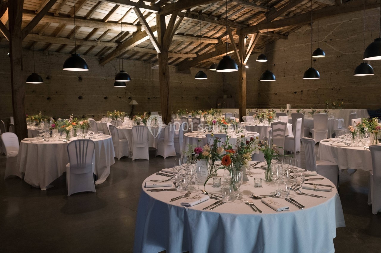 Salle de réception avec tables rondes dressées, nappes blanches, chaises couvertes et décoration florale colorée.