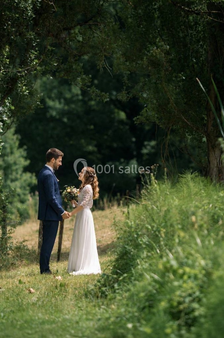 Un couple en tenue de mariage se tient la main dans un cadre naturel verdoyant sous des arbres.
