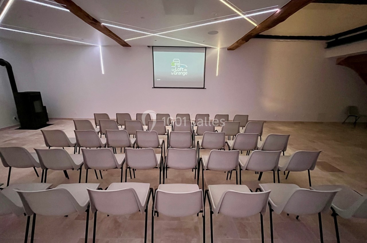 Salle de conférence avec rangées de chaises blanches alignées face à un écran de projection allumé.