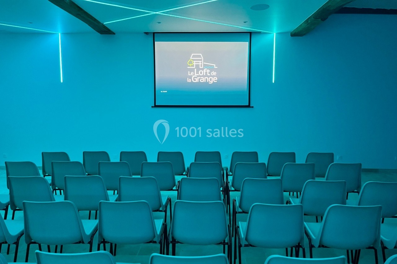 Salle de conférence moderne avec rangées de chaises vides et un écran affichant ’Le Loft de la Grange’.