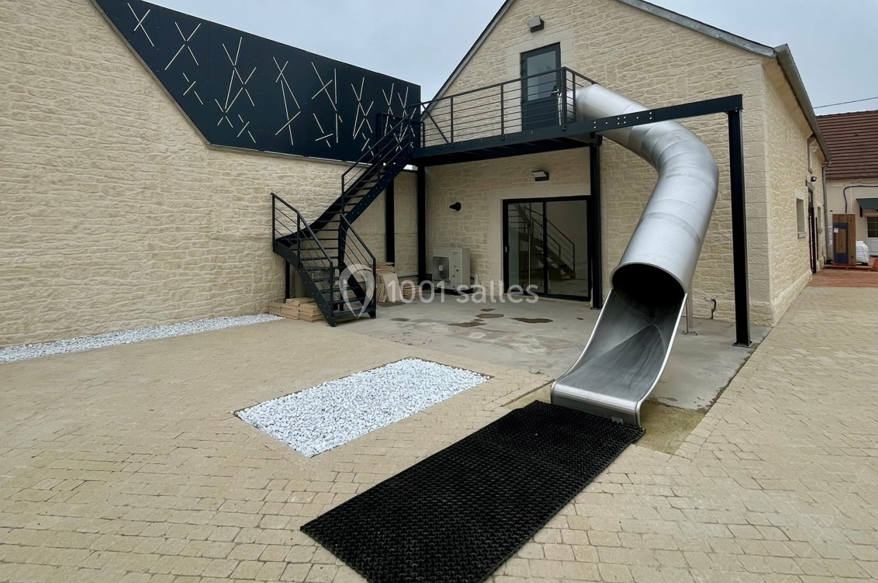 Bâtiment moderne avec un toboggan métallique descendant d'une terrasse en hauteur vers une cour pavée.