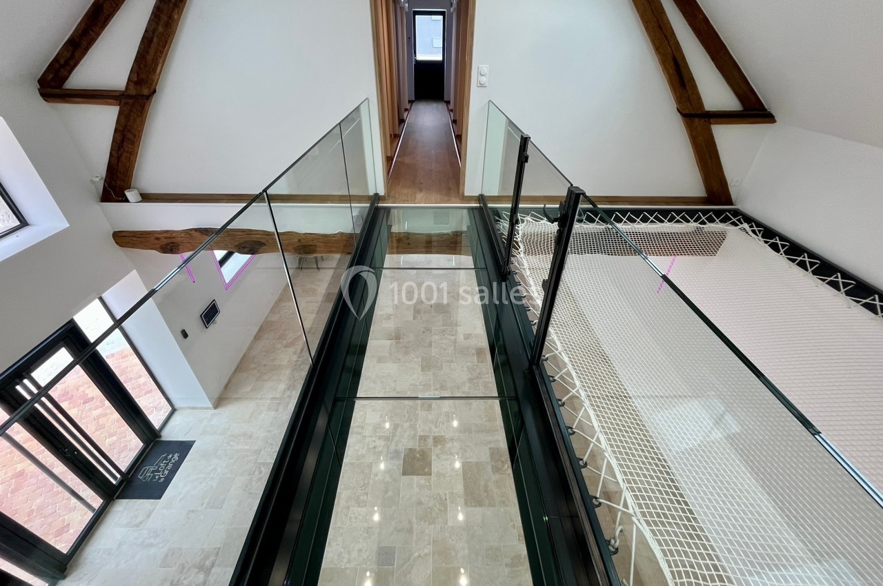 Passerelle en verre suspendue dans un intérieur moderne avec poutres apparentes et sol en pierre claire.