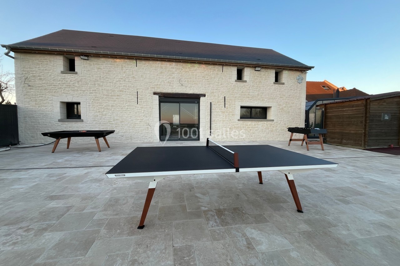 Table de ping-pong noire avec pieds en bois, placée sur une terrasse en pierre devant une maison en pierre blanche.