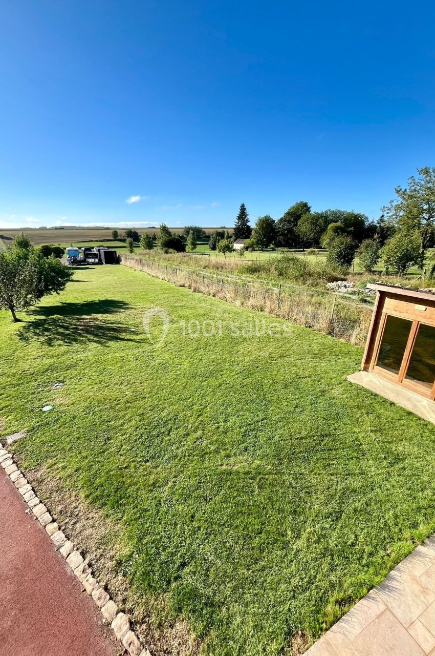 Jardin avec pelouse bien entretenue, chemin pavé, cabanon en bois et paysage rural en arrière-plan sous un ciel bleu.