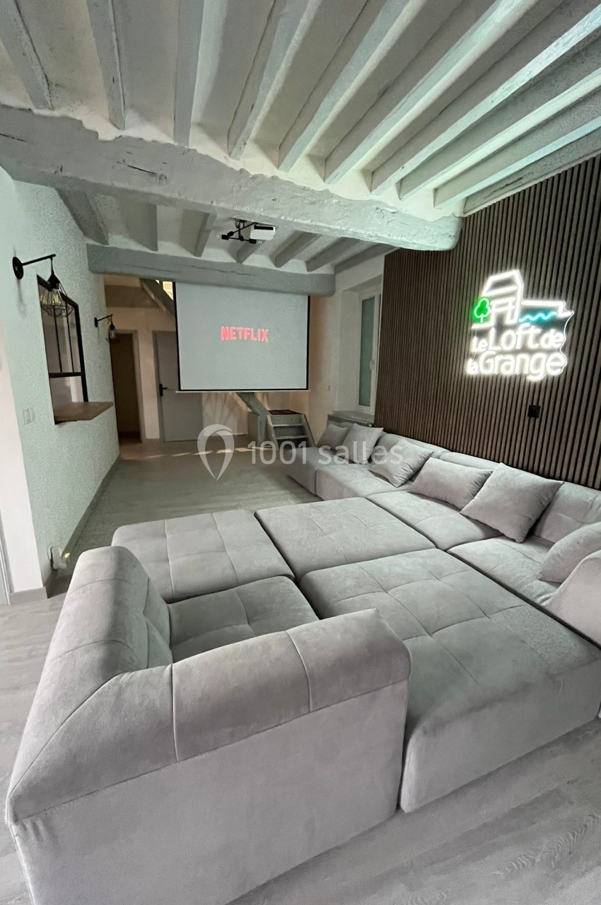 Canapé modulable gris dans un salon moderne avec écran de projection et enseigne lumineuse sur un mur en bois.