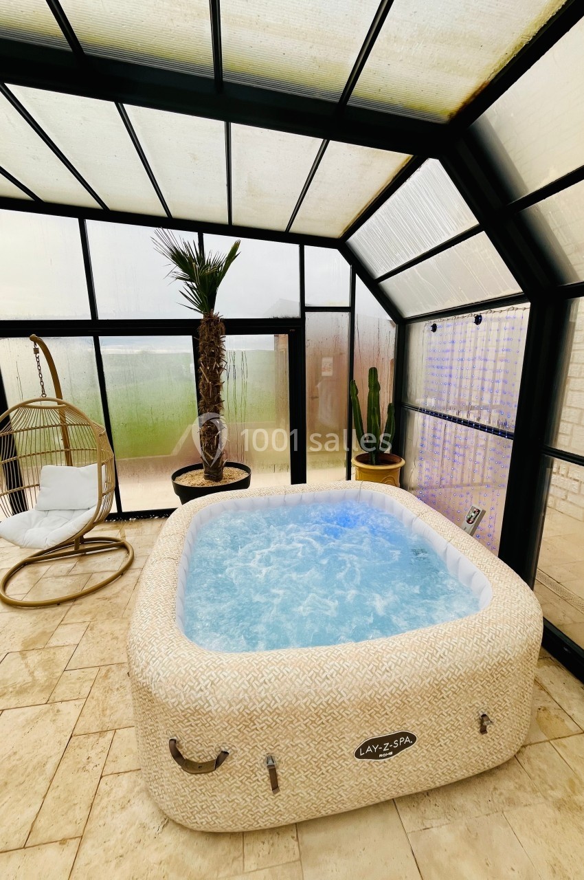Jacuzzi gonflable rempli d'eau bouillonnante dans une véranda lumineuse avec chaise suspendue, palmier et cactus.
