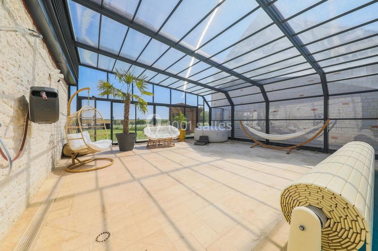 Véranda lumineuse avec piscine couverte, mobilier de détente, hamac et vue sur un jardin extérieur.