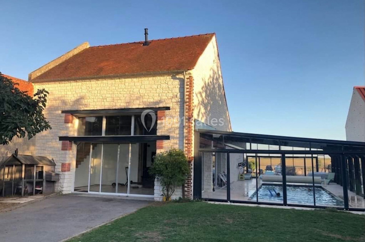 Façade d'une maison en pierre avec une véranda attenante abritant une piscine, entourée d'un jardin.