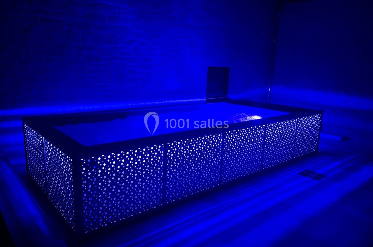 Piscine rectangulaire illuminée par une lumière bleue, entourée d'un panneau perforé dans un espace sombre.
