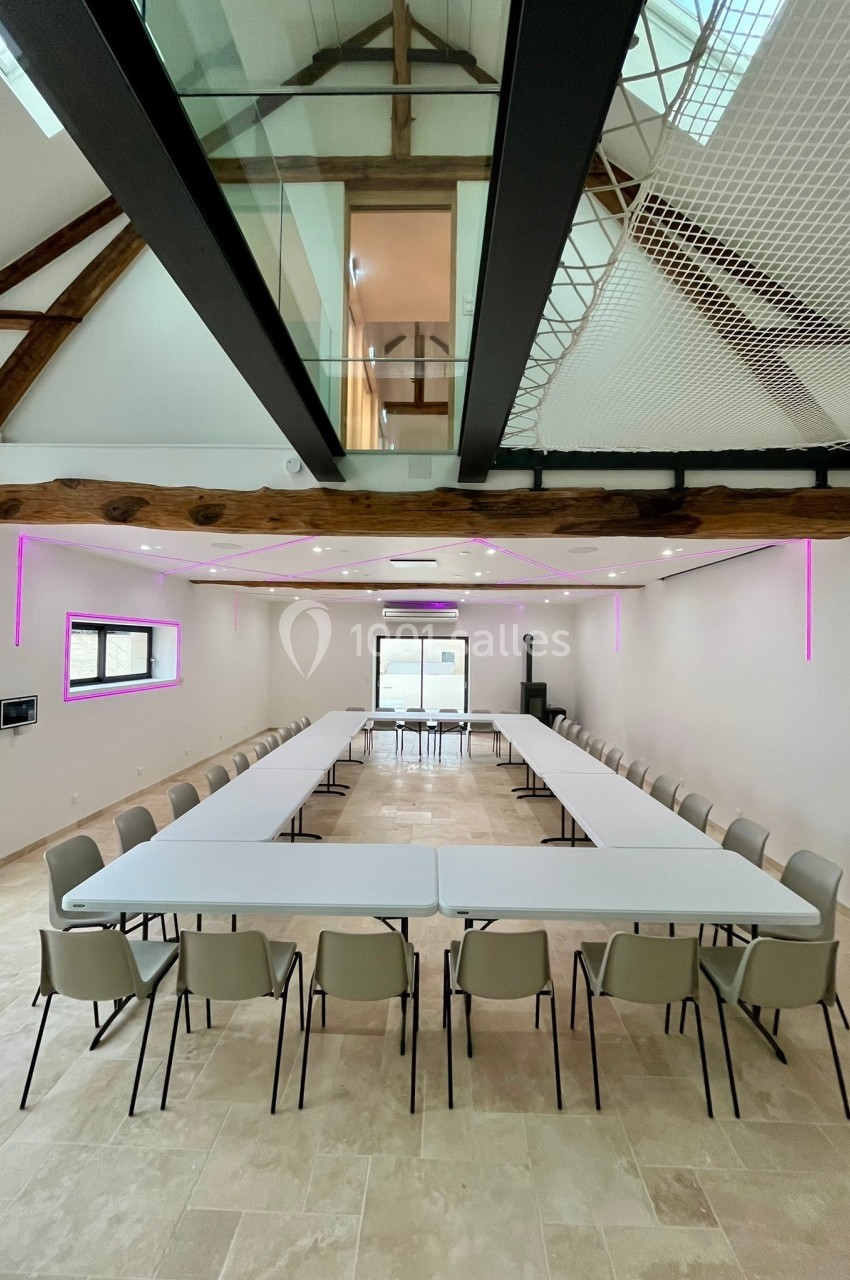 Salle de réunion lumineuse avec tables disposées en U, chaises alignées et poutres apparentes au plafond.