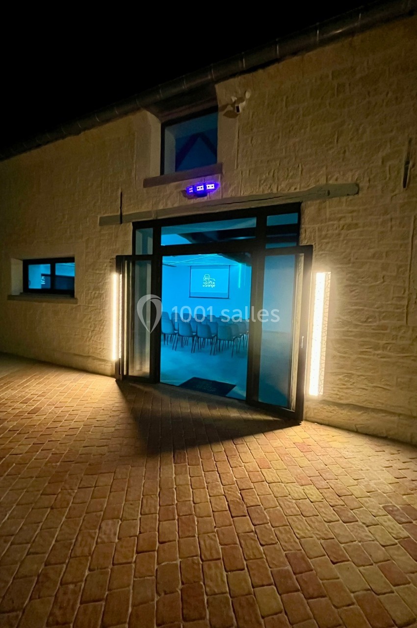 Entrée éclairée d'un bâtiment en pierre de nuit, avec une salle de conférence visible à travers les portes vitrées.