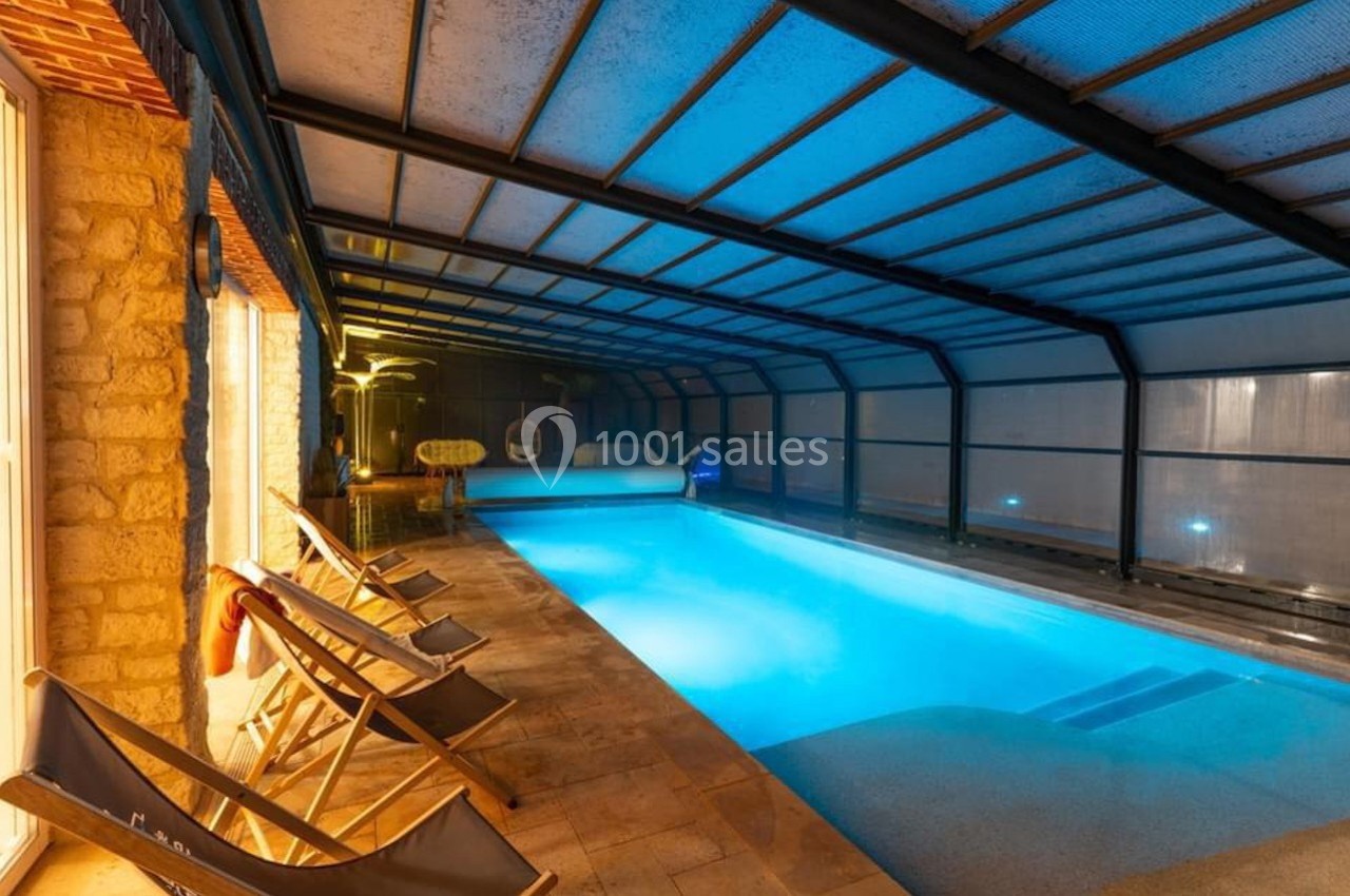 Piscine intérieure éclairée avec toit transparent, entourée de chaises longues et d'un sol en pierre.