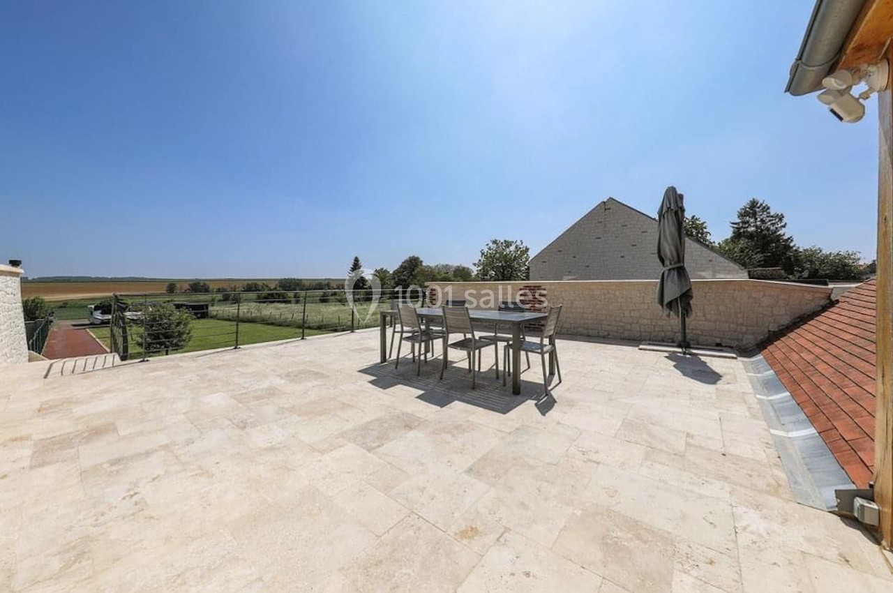 Terrasse spacieuse en pierre avec table, chaises et parasol, offrant une vue dégagée sur la campagne environnante.