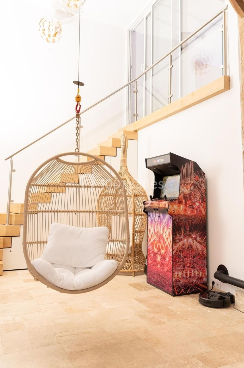Fauteuil suspendu en rotin avec coussin blanc près d'un escalier moderne et d'une borne d'arcade colorée.