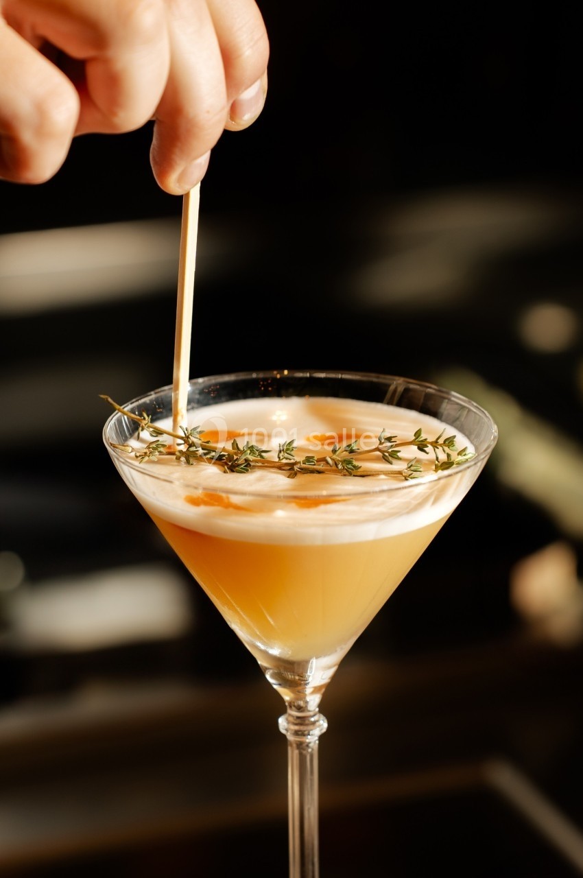 Un cocktail jaune décoré de thym et mousse blanche, avec une main ajustant une brochette en bois.