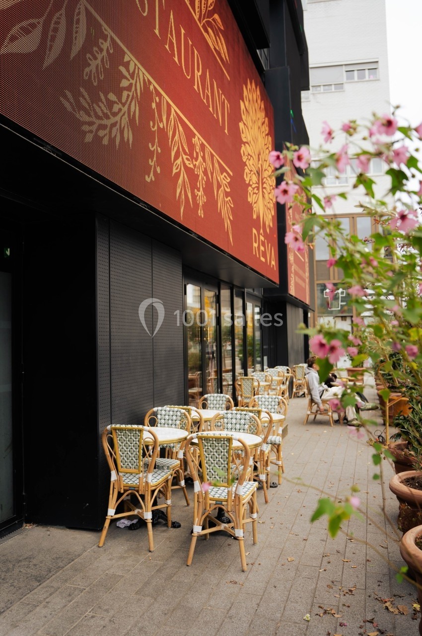 Terrasse d'un restaurant avec des chaises en rotin, entourée de plantes en pot et située près d'un bâtiment moderne.