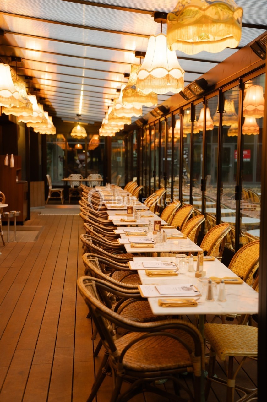 Salle de restaurant avec tables alignées, chaises en rotin et éclairage chaleureux sous une verrière.