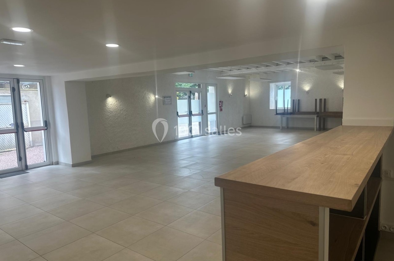 Salle lumineuse avec carrelage clair, comptoir en bois à droite, grandes fenêtres et portes vitrées donnant sur l'extérieur.