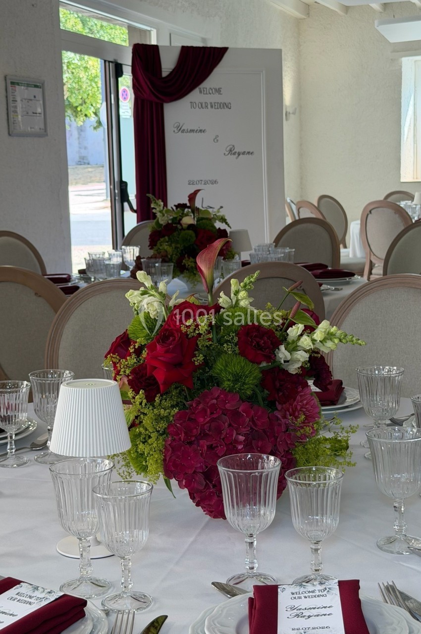 Salle de réception décorée avec des tables dressées, des fleurs rouges et blanches, et un panneau de bienvenue au fond.
