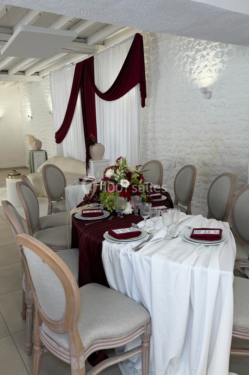 Table élégamment dressée avec nappes blanches et bordeaux, décor floral central et chaises rembourrées dans une salle…