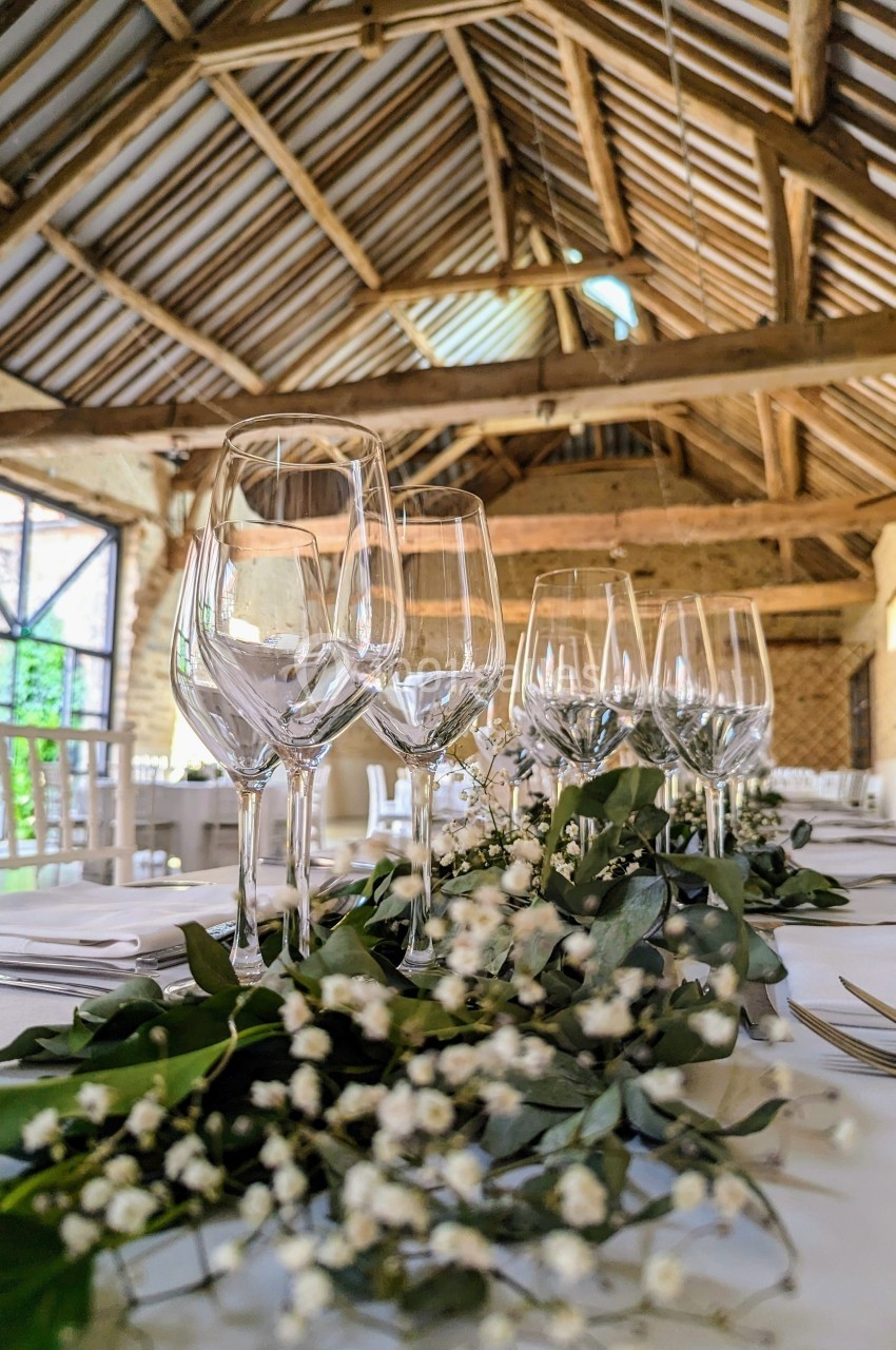 Table décorée avec des verres à pied, couverts et guirlande végétale dans une salle en pierre avec charpente apparente.