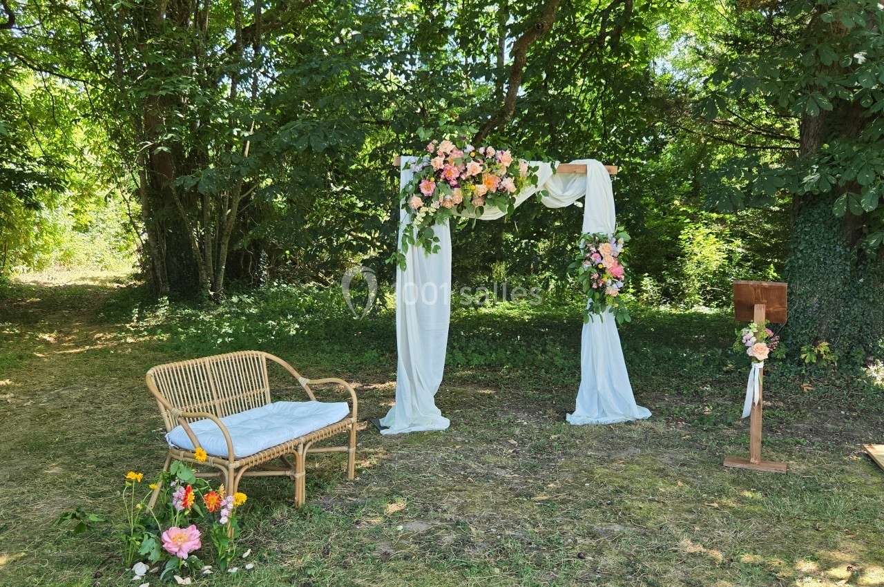 Arche de mariage décorée de fleurs et de voilages blancs, avec un banc en rotin et des fleurs au sol, dans un jardin ombragé.