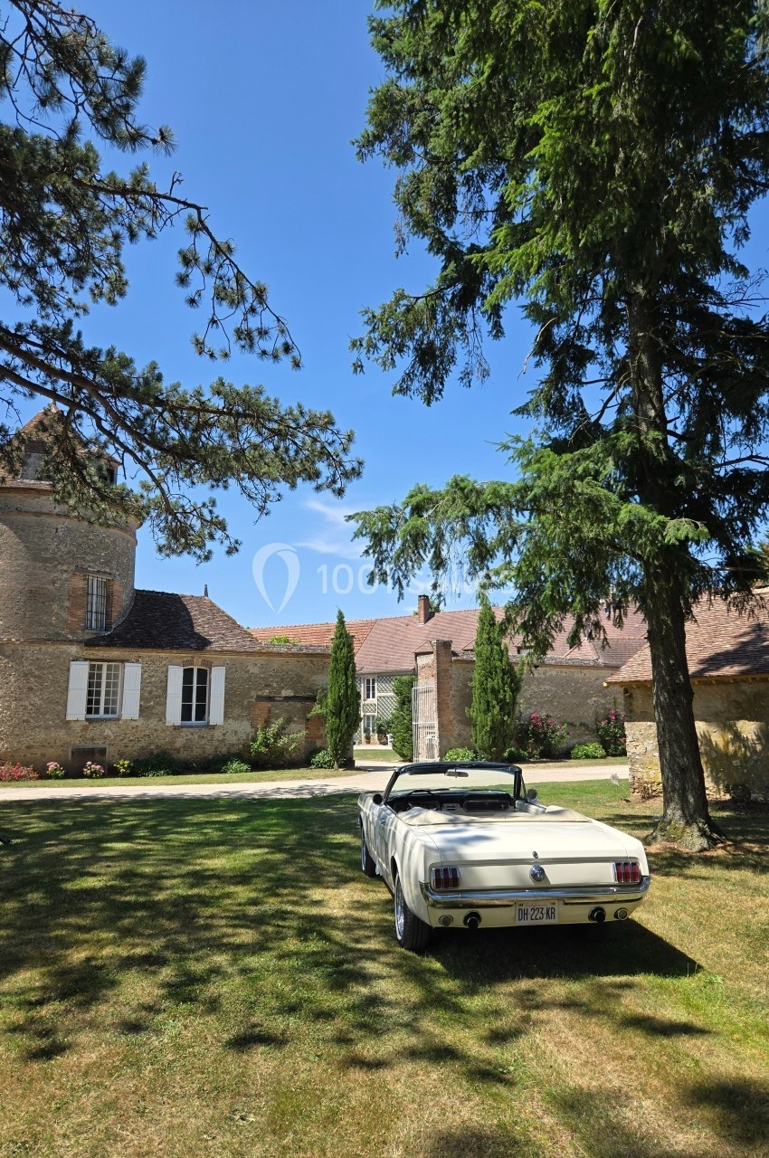 Voiture décapotable blanche garée sur une pelouse devant un bâtiment en pierre avec des arbres et un ciel bleu.