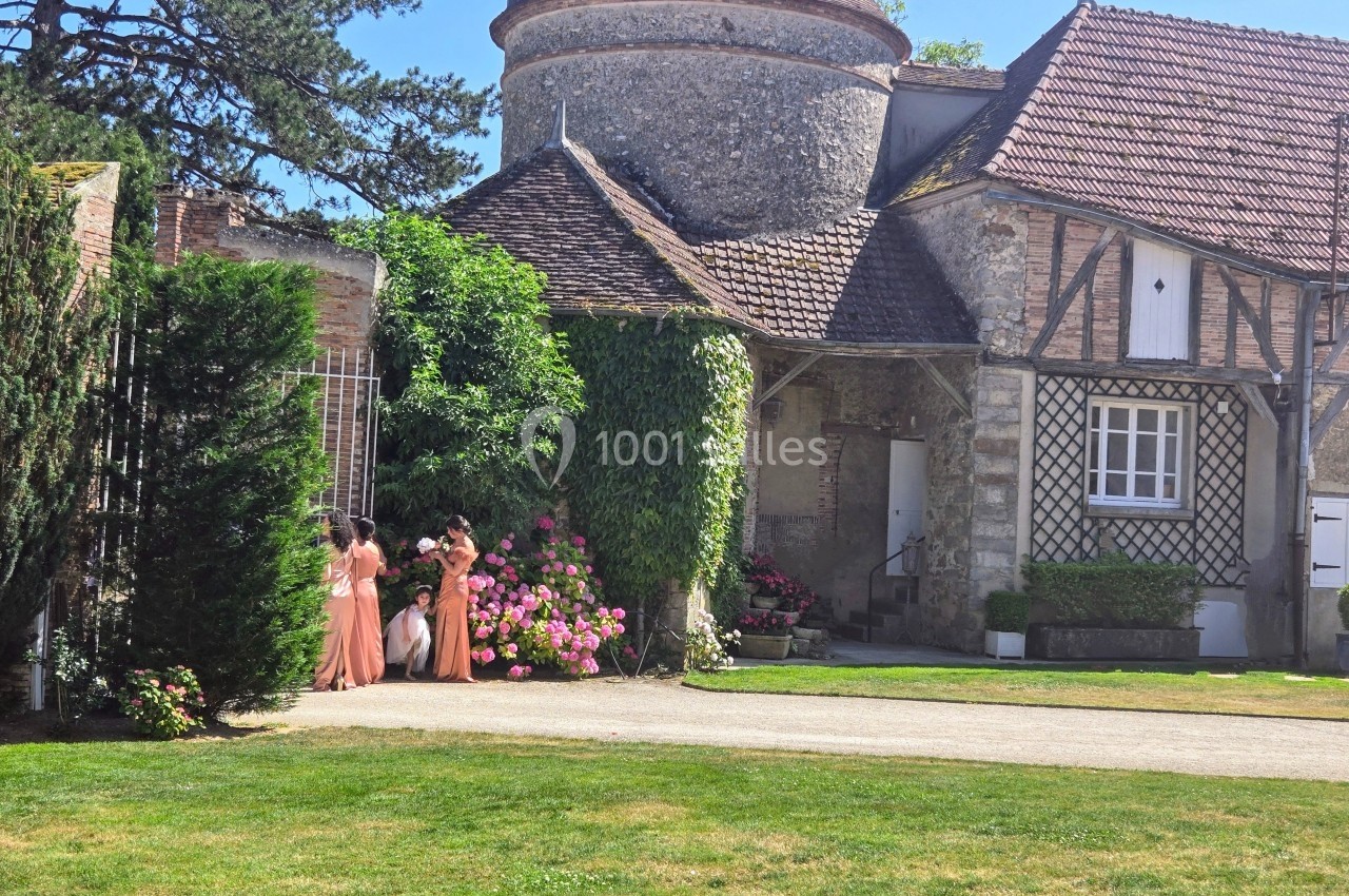 Deux femmes en robes roses marchent près d'un bâtiment en pierre entouré de fleurs et de verdure.
