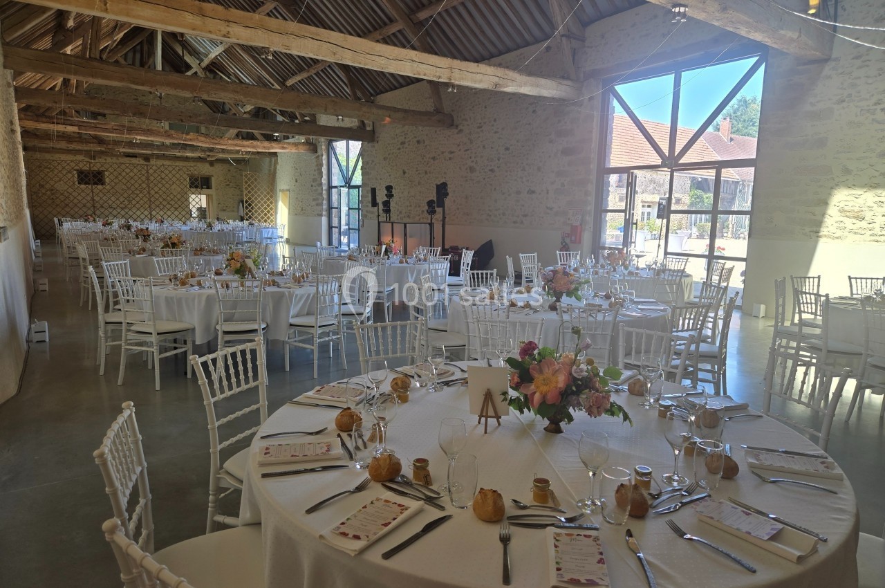 Salle de réception lumineuse avec tables rondes décorées de fleurs et chaises blanches, prête pour un événement.